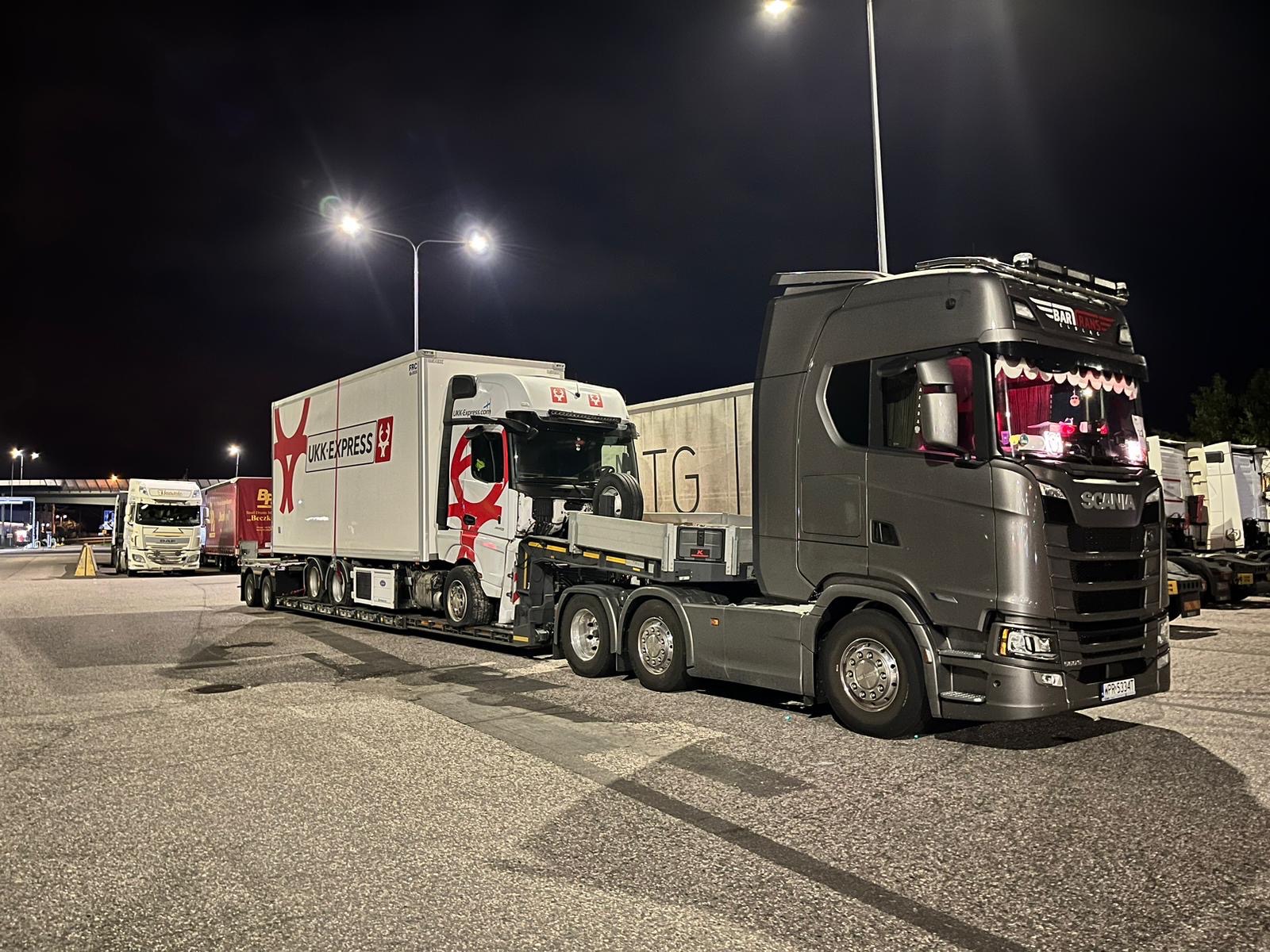 Nocny widok na parking z zaparkowanymi ciężarówkami, w tym szarą Scanię z ozdobnymi zasłonkami w kabinie i białą ciężarówką UKK Express z czerwonym logo, stojące pod latarniami.