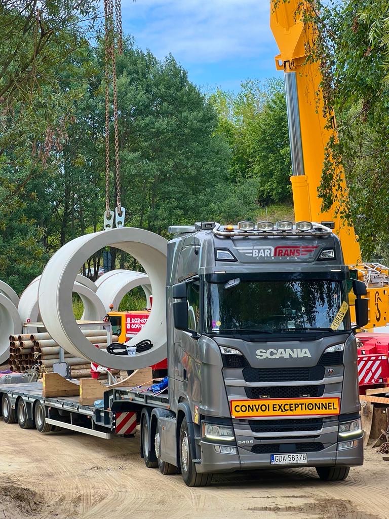 Szary ciągnik siodłowy Scania z naczepą przewożący betonowe kręgi, z żółtym dźwigiem w tle i tablicą 'Convoi Exceptionnel' z przodu kabiny.