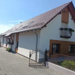 Odnowiony dom jednorodzinny z białą elewacją i brązowymi akcentami, widoczny z zewnątrz, z otwartą kutą bramą i ozdobnym balkonem z kwiatami.