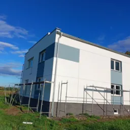 Dwukondygnacyjny dom z częściowo otynkowaną elewacją w odcieniach bieli i szarości, otoczony rusztowaniami na tle błękitnego nieba z lekkimi obłokami.