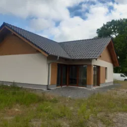 Nowoczesny parterowy dom z beżową elewacją, grafitową dachówką i drewnianymi elementami wykończeniowymi, widziany z perspektywy trawnika z dzikimi kwiatami; w tle zielone drzewa i maszt...