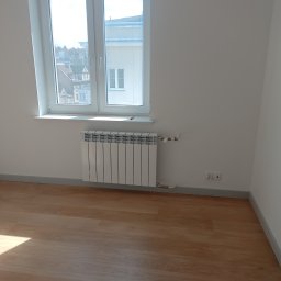 A-Z Hydraulik - Biały, aluminiowy grzejnik żeberkowy zamontowany pod oknem w nowo wykończonym pokoju z parkietem i szarą listwą przypodłogową.