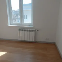 Biały, aluminiowy grzejnik żeberkowy zamontowany pod oknem w nowo wykończonym pokoju z parkietem i szarą listwą przypodłogową.