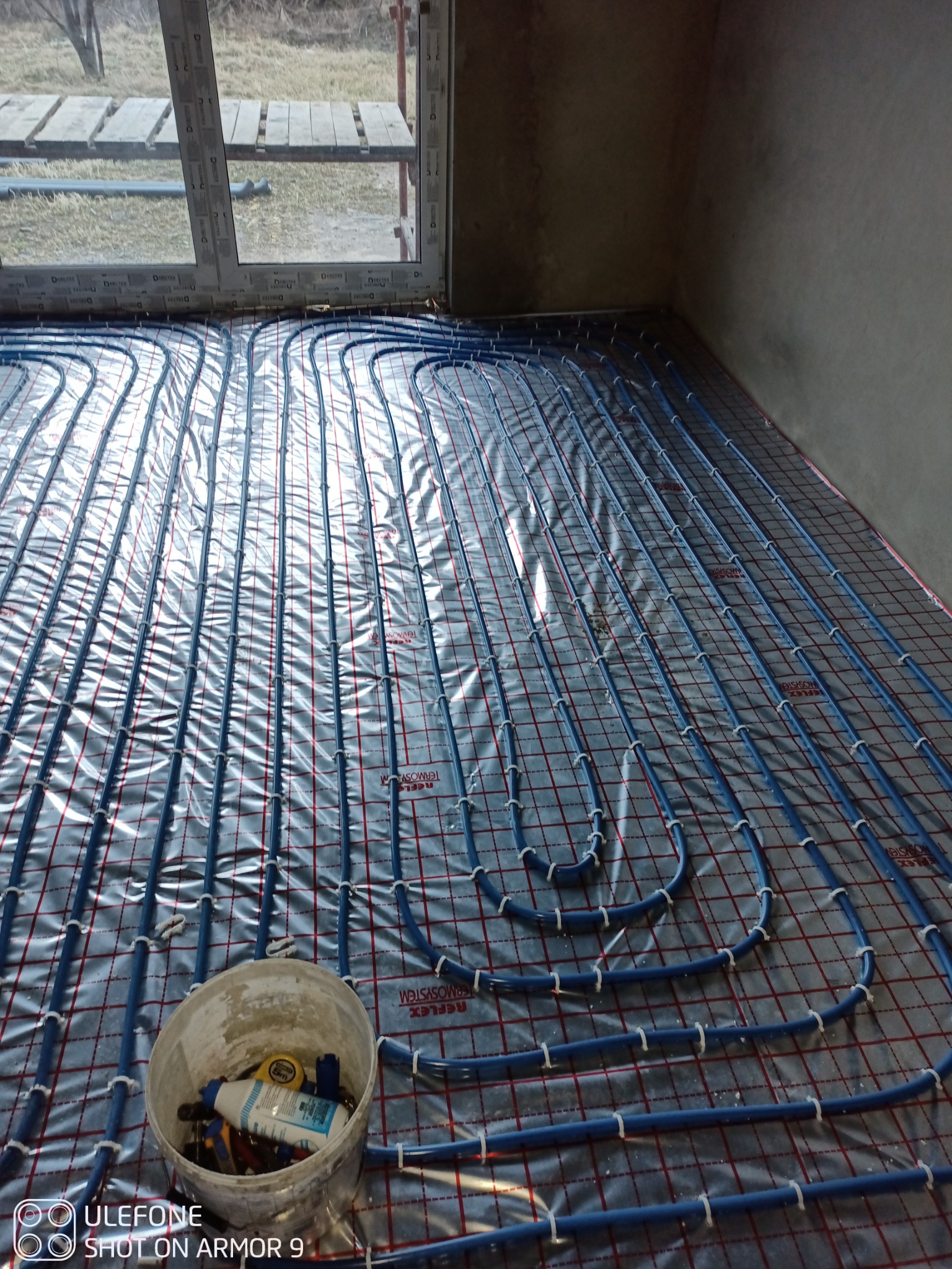 Instalacja ogrzewania podłogowego: niebieskie rurki ułożone na folii izolacyjnej z widocznym wiaderkiem z narzędziami w rogu pomieszczenia.