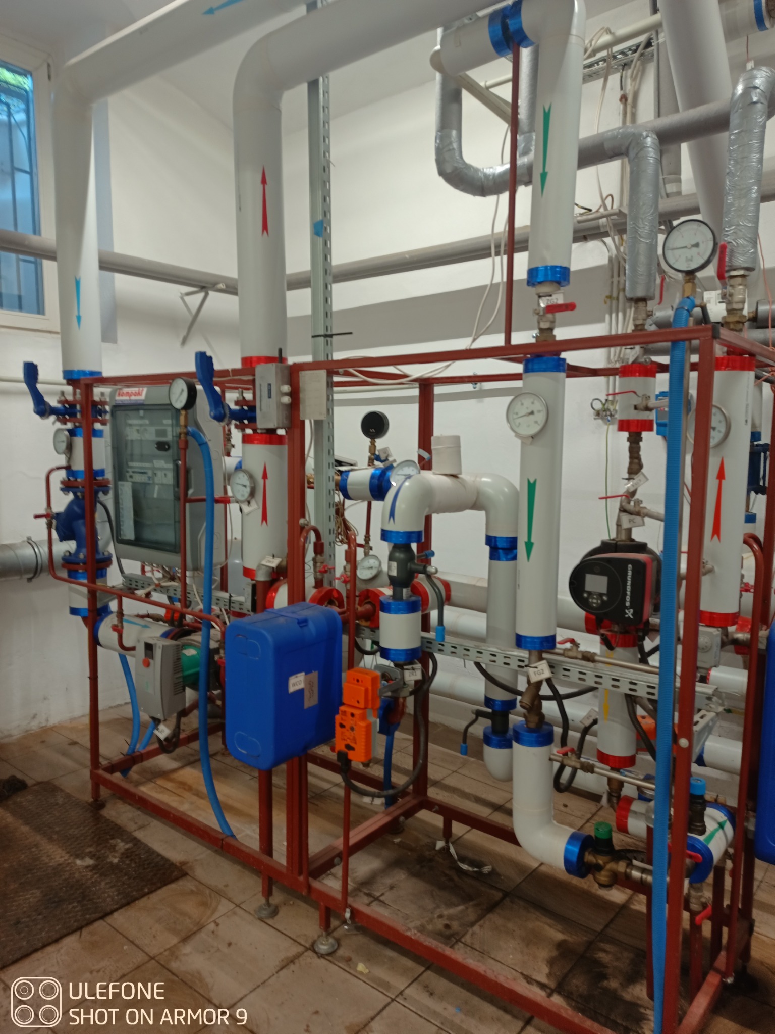 Skomplikowany system rur i zaworów w kotłowni, z widocznymi manometrami, pompą obiegową Grundfos i elementami instalacji elektrycznej, całość osadzona na metalowej konstrukcji.