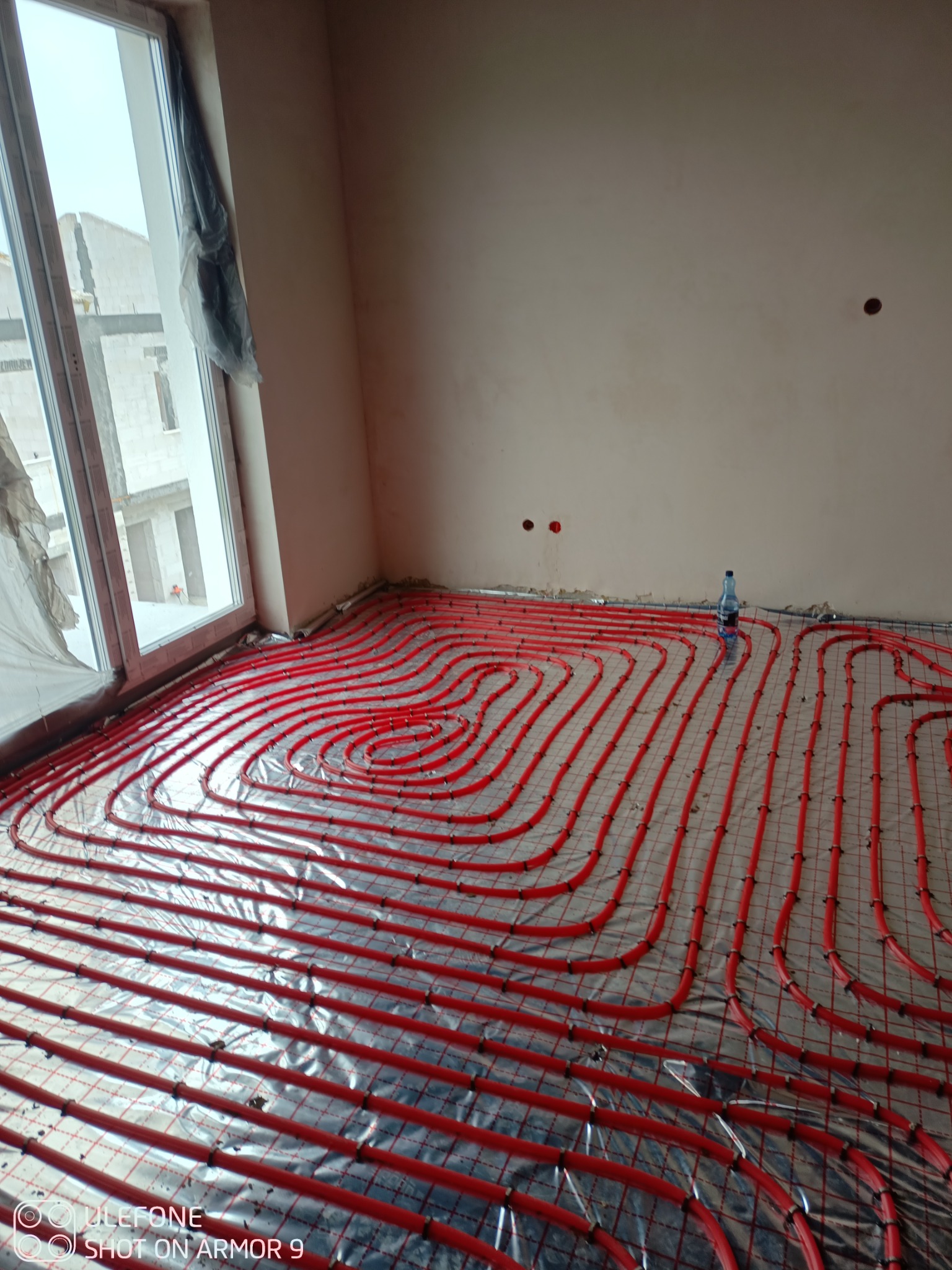 Instalacja czerwonych rur ogrzewania podłogowego na folii aluminiowej, widoczne mocowania, w tle okno z folią ochronną i butelka wody.