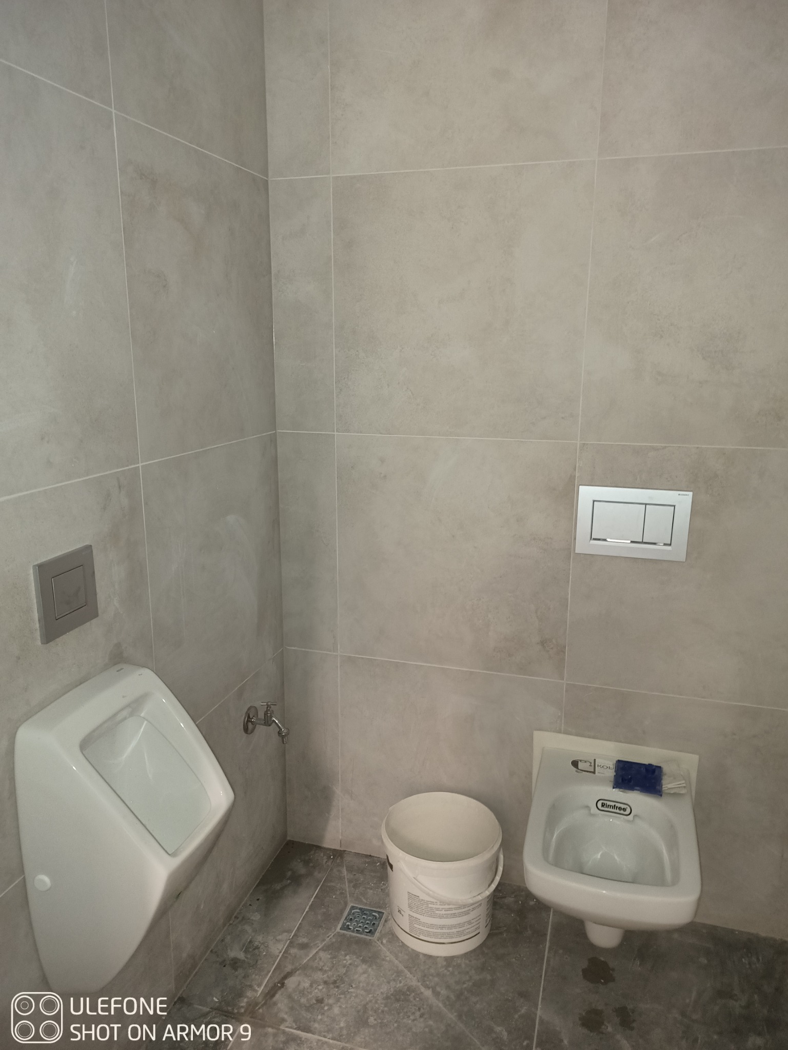 Ukończona łazienka z szarymi płytkami, pisuar i umywalka dla dzieci, widoczny biały plastikowy pojemnik.