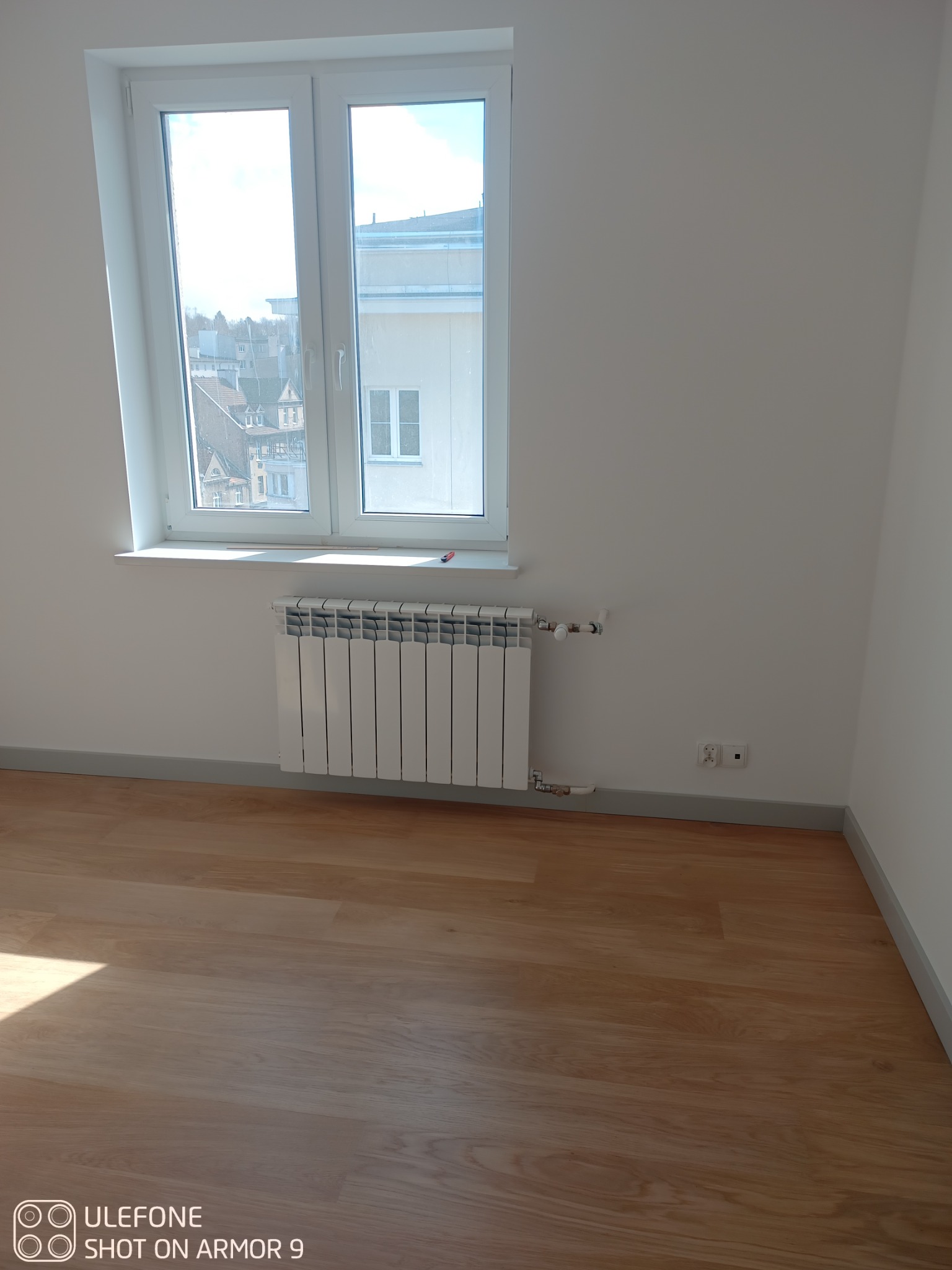 Biały, aluminiowy grzejnik żeberkowy zamontowany pod oknem w nowo wykończonym pokoju z parkietem i szarą listwą przypodłogową.
