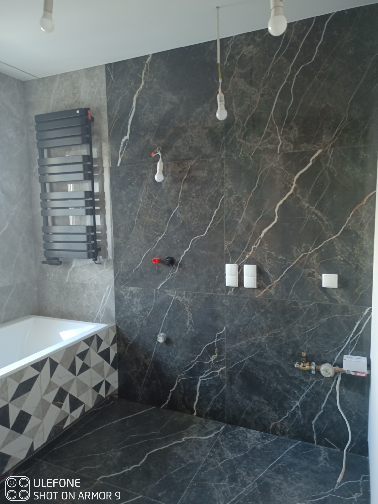 Częściowo wykończona łazienka z czarno-białymi marmurkowymi płytkami, grzejnikiem drabinkowym, białą wanną z geometrycznym wzorem na obudowie, widoczne elementy instalacji hydraulicznej...