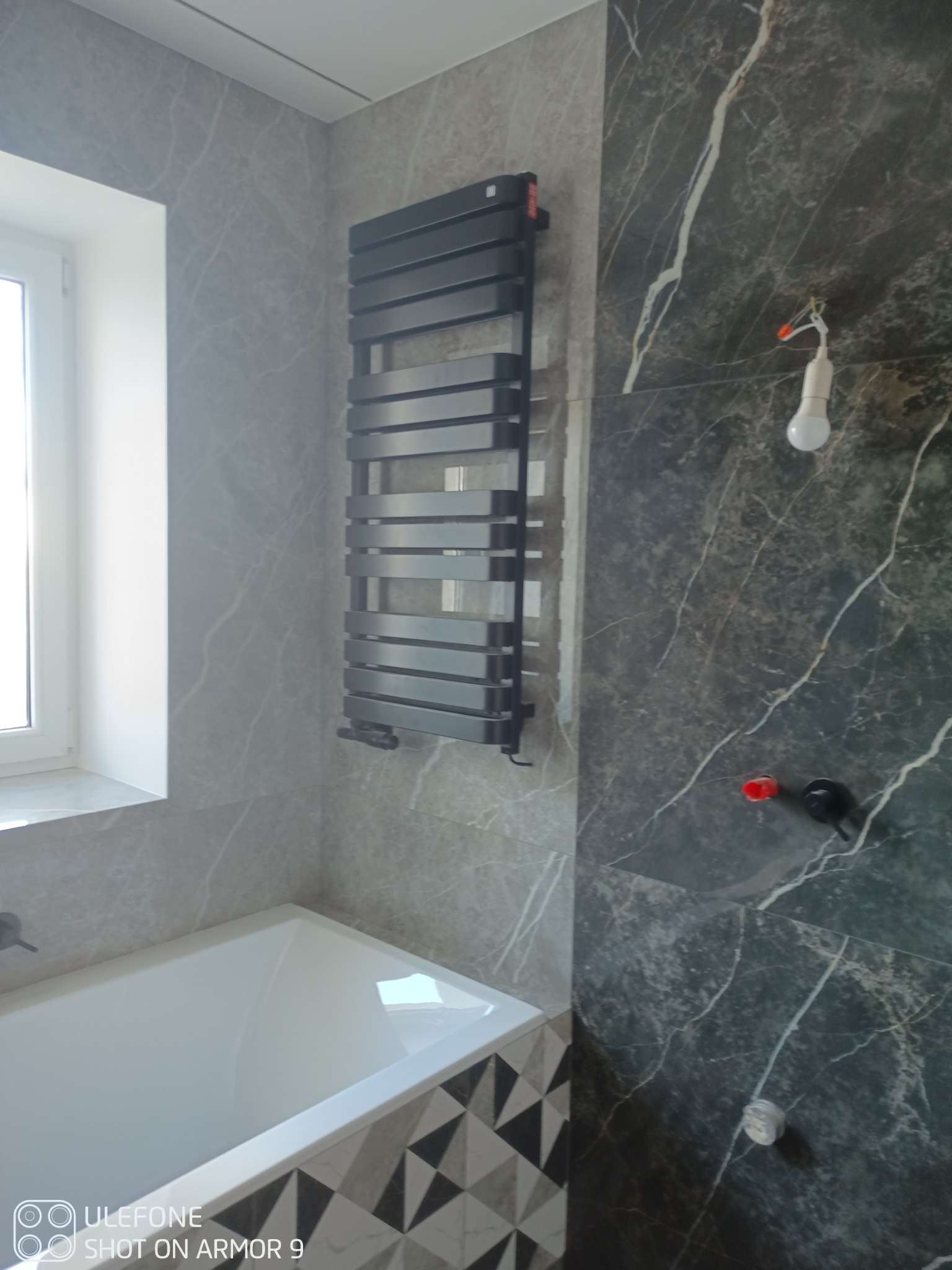 Łazienka w trakcie remontu: biała wanna z geometrycznym wzorem na obudowie, szary grzejnik drabinkowy na ścianie z płytek imitujących marmur, widoczne przewody i żarówka.