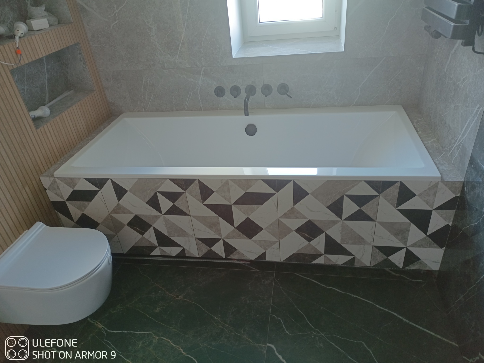 Wykończona łazienka z białą wanną obudowaną płytkami w geometryczny wzór, toaleta typu Geberit z widoczną spłuczką, szare marmurowe płytki ścienne i podłogowe, okno dachowe.