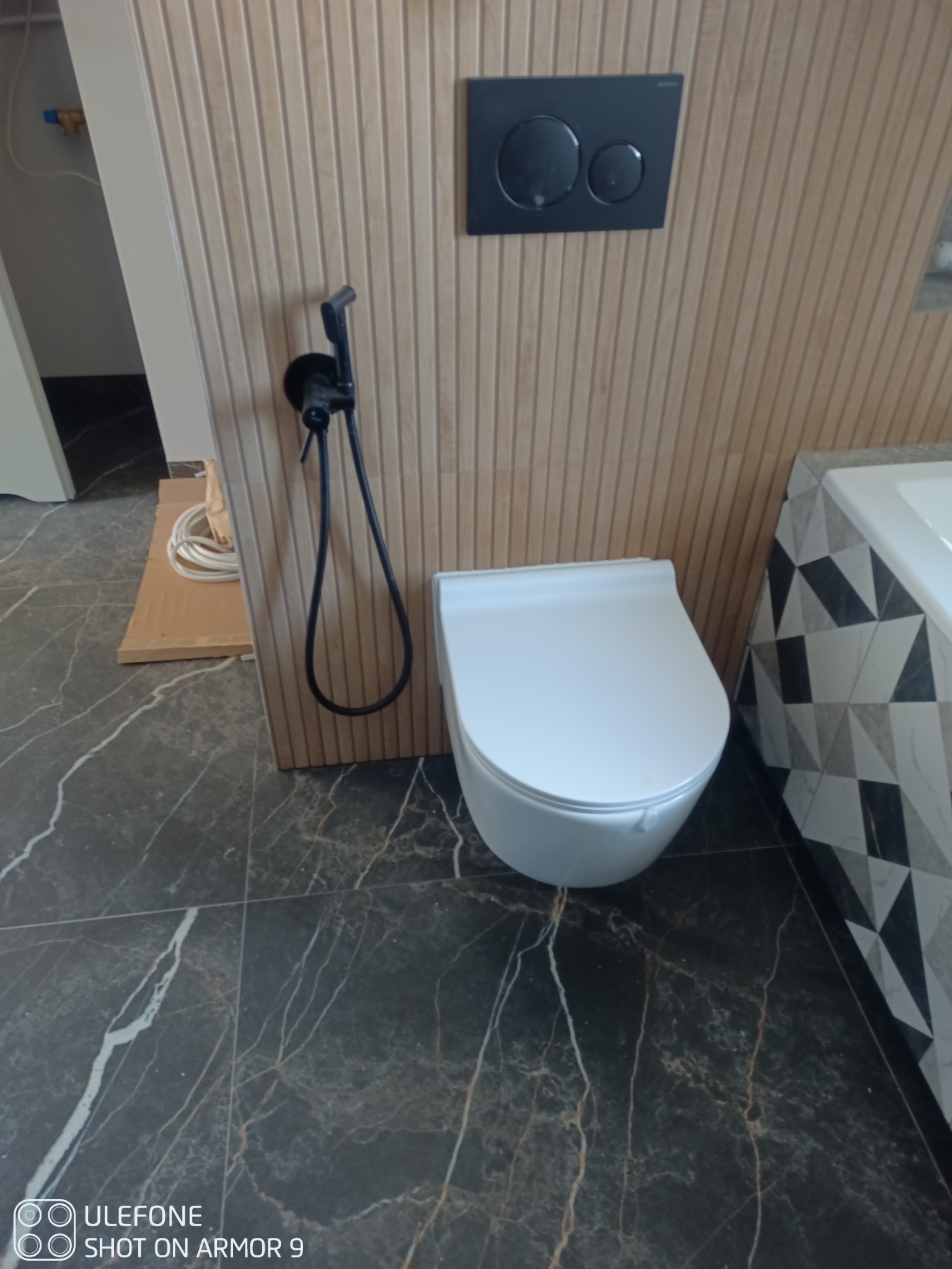 Nowoczesna toaleta podwieszana z bidetką, zamontowana na ścianie wykończonej drewnopodobnymi lamelami, z czarnym panelem sterującym spłukiwaniem. Na podłodze ciemne płytki imitujące marmur.