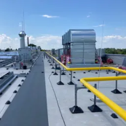 Instalacje wentylacyjne i systemy zabezpieczeń na dachu budynku przemysłowego, widoczne elementy instalacji odgromowej, żółta balustrada ochronna i panele solarne w tle.
