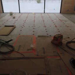 Przygotowanie do układania płytek podłogowych w nowym domu z dużym oknem na zimowy krajobraz. Widoczne narzędzia, poziomowanie i kartony ochronne Marazzi.