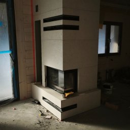 Wykończony kominek z jasnego kamienia z czarnymi akcentami w surowym wnętrzu. Widoczne narzędzia i resztki materiałów budowlanych na podłodze. Częściowo zakryte okno.