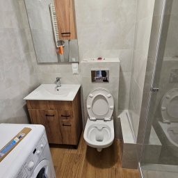 Wykończona łazienka z drewnopodobną podłogą, umywalką z szafką, toaletą podwieszaną i kabiną prysznicową. Uwagę zwraca drewniana szafka z lustrem nad umywalką.