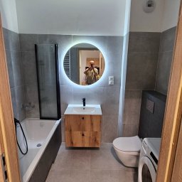Usługi budowlane - Nowoczesna łazienka z szarymi płytkami, okrągłym lustrem z podświetleniem LED, drewnianą szafką pod umywalką, wanną z parawanem i pralką. Widok zza futryny.