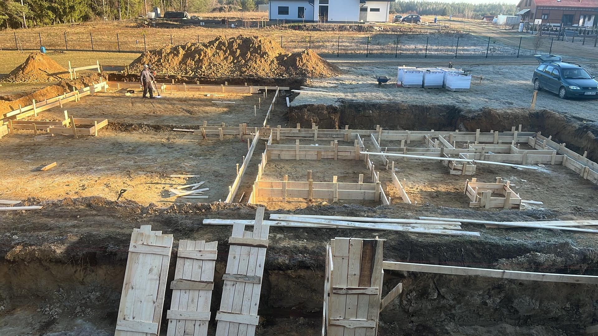 Fundamenty domu jednorodzinnego w trakcie budowy: wykopane rowy, drewniane szalunki, ziemia, w tle domy i drzewa. Widoczny pracownik budowlany.