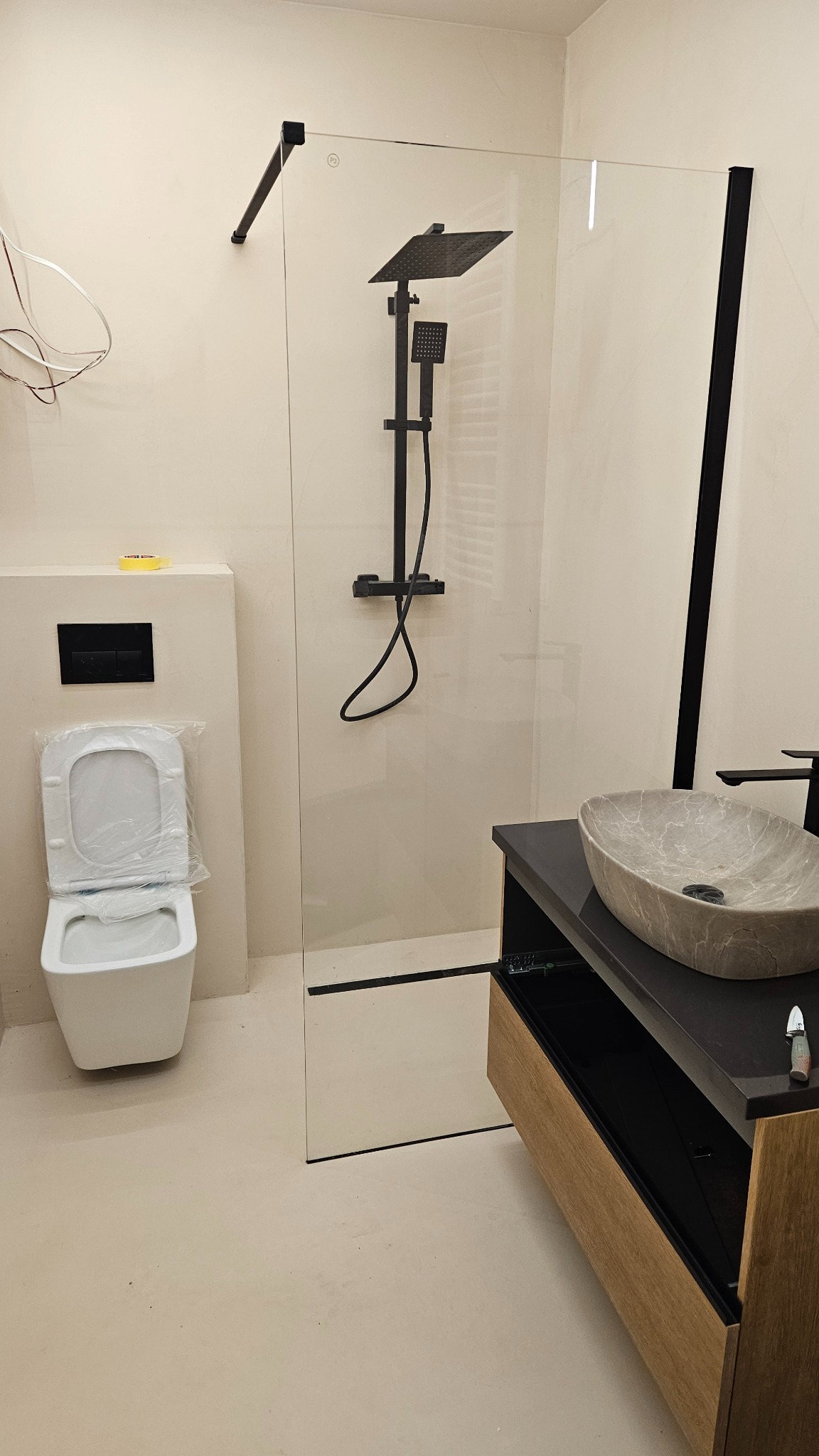 Nowoczesna łazienka w trakcie wykończenia: kabina prysznicowa typu walk-in z czarną armaturą, umywalka nablatowa z szarego kamienia i podwieszana toaleta z folią ochronną.