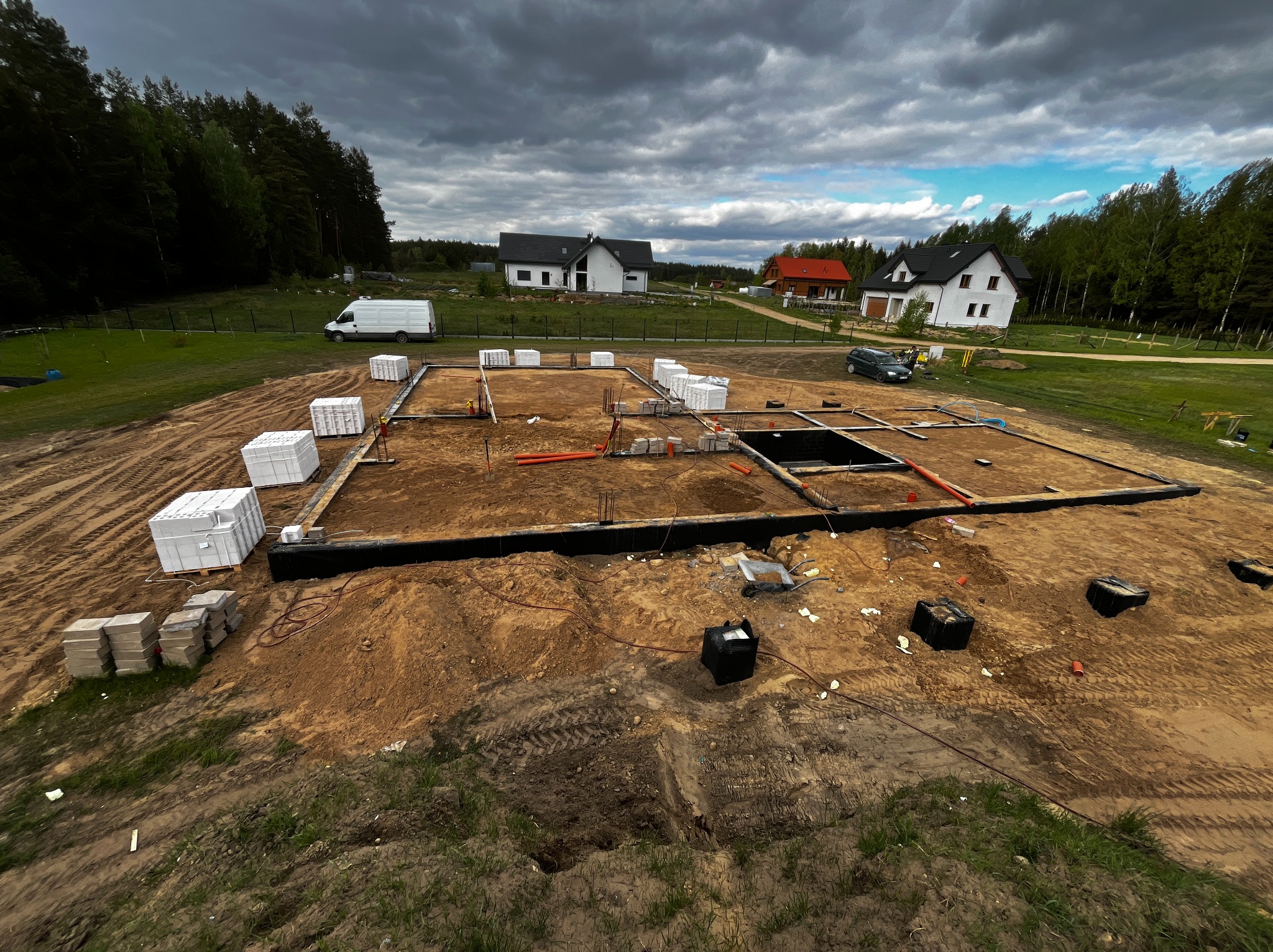 Fundamenty domu jednorodzinnego w trakcie budowy, widok z góry. Ułożone bloczki, instalacje, teren przygotowany pod wylewkę. W tle zabudowa jednorodzinna i las.