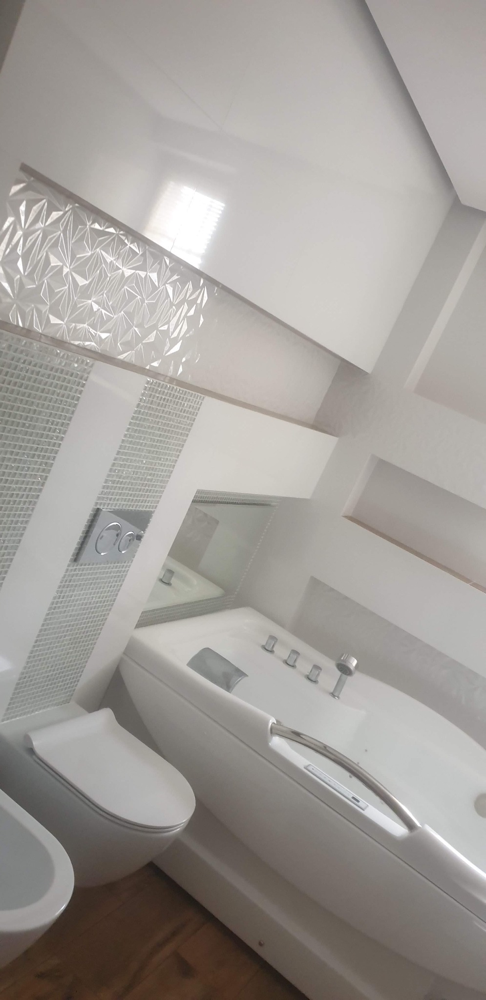 Biała, nowoczesna łazienka z wanną, toaletą i bidetem. Ściany wykończone białymi płytkami z dekoracyjnym wzorem. Uwagę zwraca minimalistyczny design i jasne oświetlenie.