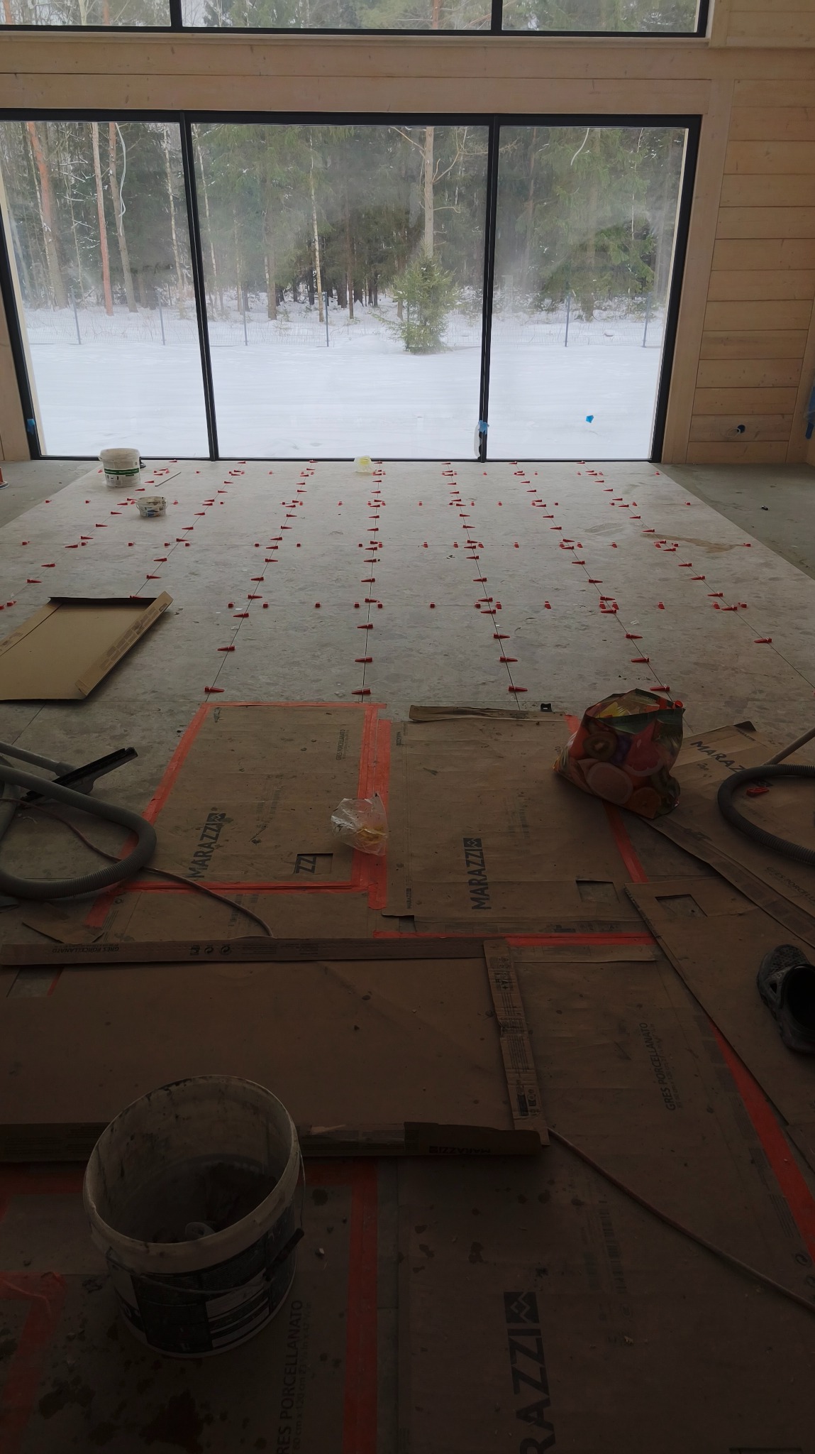 Przygotowanie do układania płytek podłogowych w nowym domu z dużym oknem na zimowy krajobraz. Widoczne narzędzia, poziomowanie i kartony ochronne Marazzi.