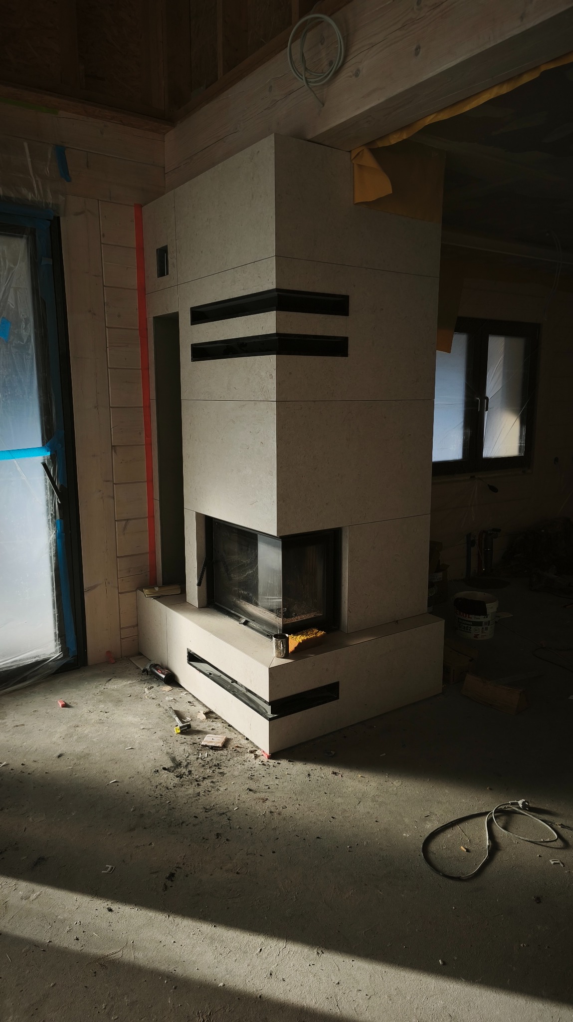 Wykończony kominek z jasnego kamienia z czarnymi akcentami w surowym wnętrzu. Widoczne narzędzia i resztki materiałów budowlanych na podłodze. Częściowo zakryte okno.