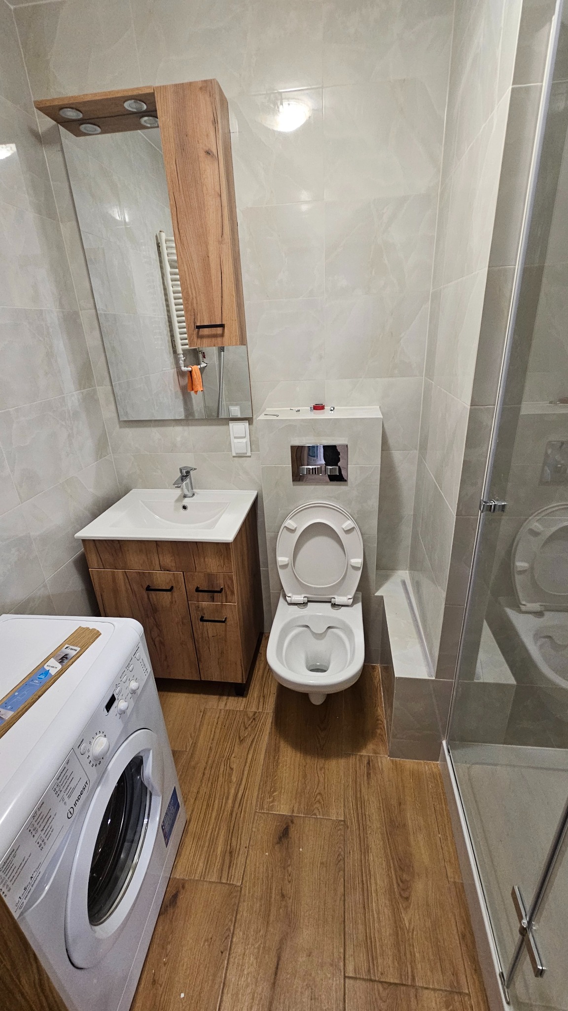 Wykończona łazienka z drewnopodobną podłogą, umywalką z szafką, toaletą podwieszaną i kabiną prysznicową. Uwagę zwraca drewniana szafka z lustrem nad umywalką.