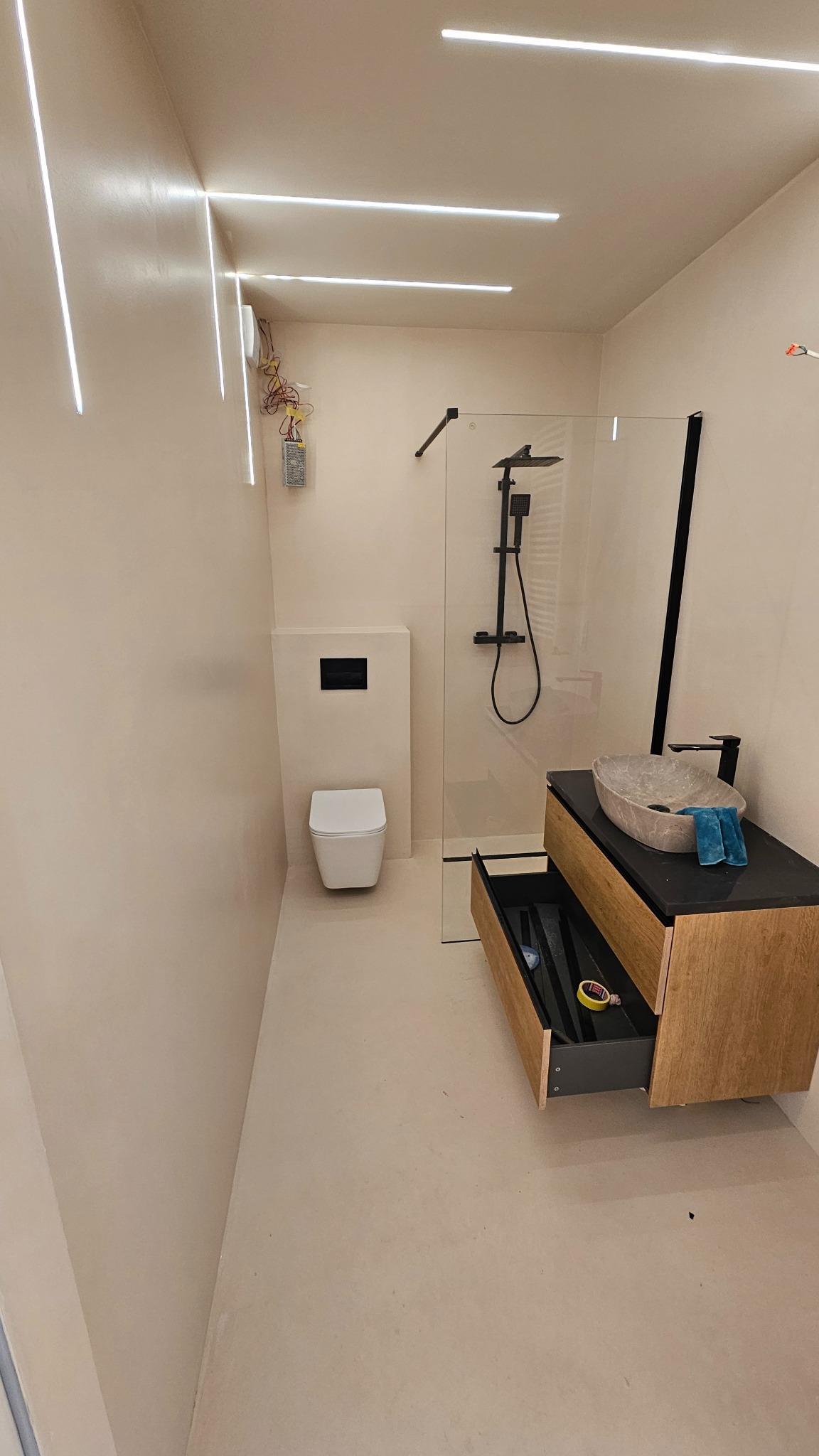 Nowoczesna łazienka z liniowym oświetleniem LED, kabiną prysznicową typu walk-in, umywalką nablatową i szafką z otwartą szufladą. Minimalistyczny design, beżowe ściany.