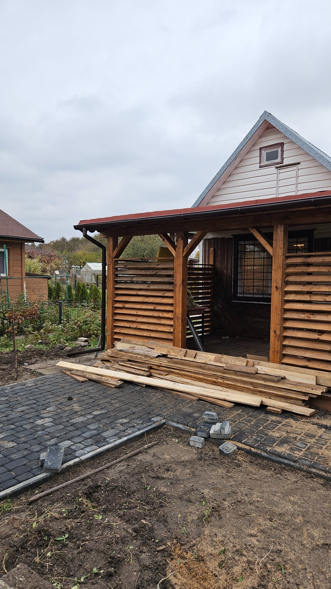 Drewniana konstrukcja z lamelami w ogrodzie, częściowo wykończona kostka brukowa. Widoczne deski i elementy konstrukcyjne. Prace w toku, pochmurne niebo.
