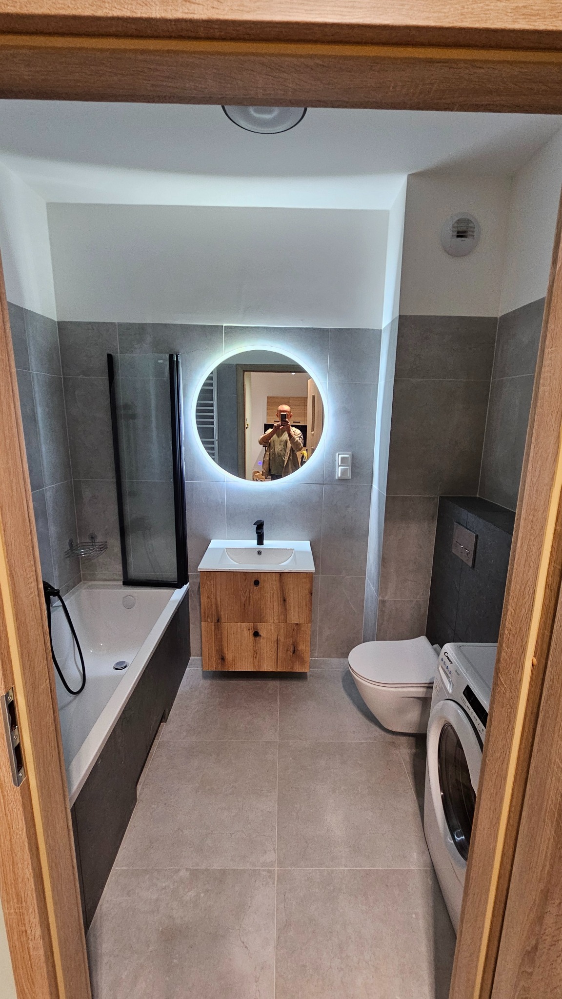 Nowoczesna łazienka z szarymi płytkami, okrągłym lustrem z podświetleniem LED, drewnianą szafką pod umywalką, wanną z parawanem i pralką. Widok zza futryny.