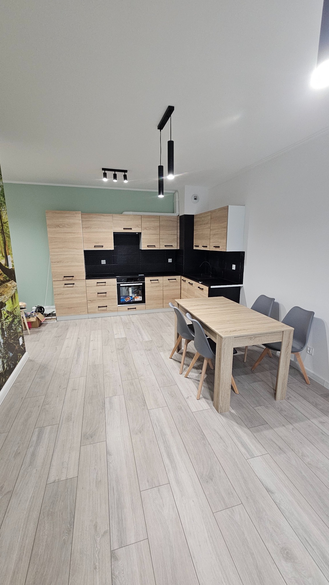 Jasna kuchnia z drewnianymi szafkami i czarnym backsplashiem, stół z szarymi krzesłami, panele podłogowe imitujące drewno. Ściana z fototapetą z motywem leśnym.