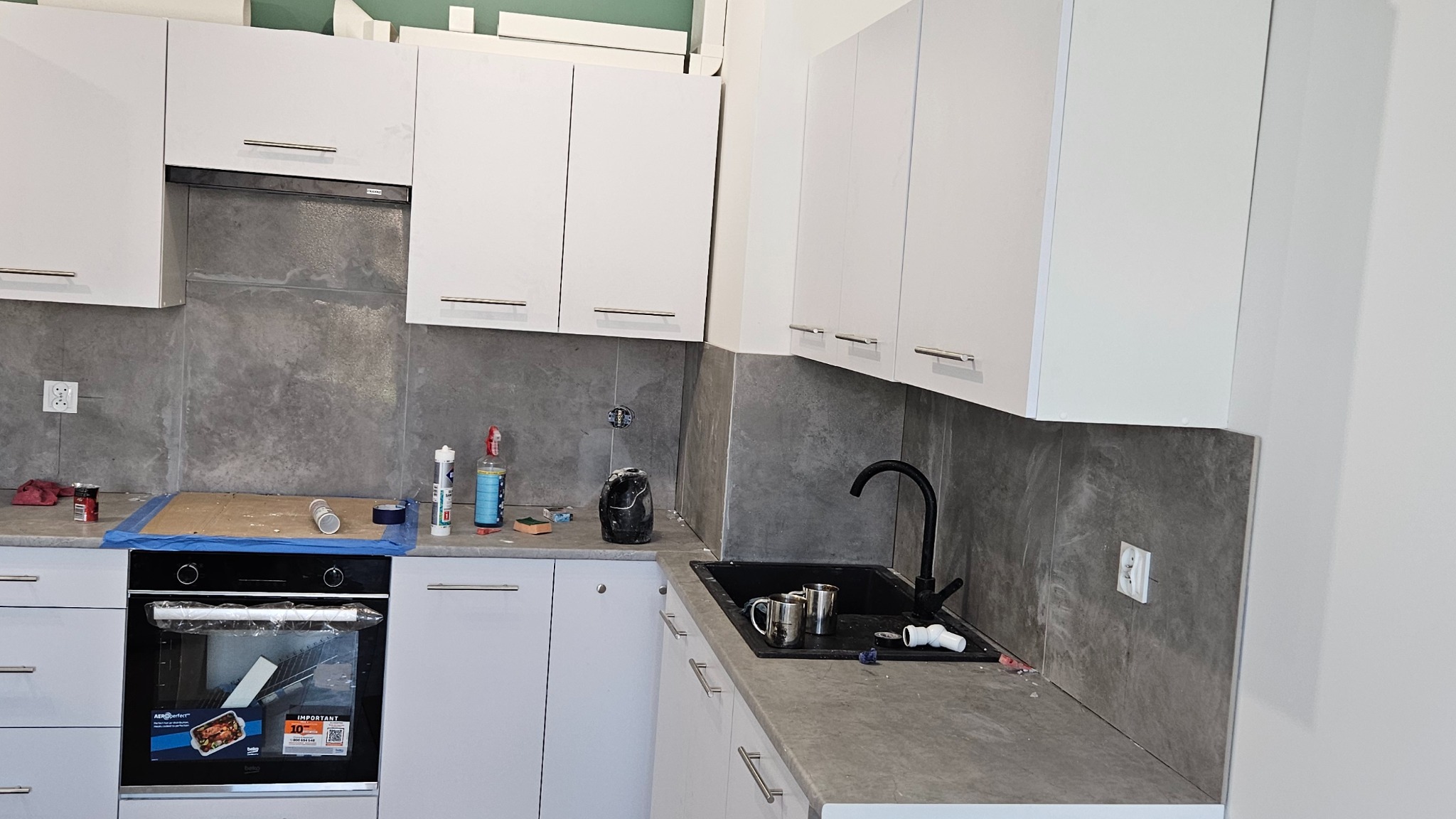 Nowoczesna kuchnia z białymi szafkami i szarymi płytkami na ścianie. Widoczny czarny zlew i bateria. Na blacie akcesoria kuchenne i drobne narzędzia.