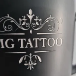 Czarny kubek z wygrawerowanym logo 'MG TATTOO' i ozdobnymi elementami graficznymi w stylu vintage.