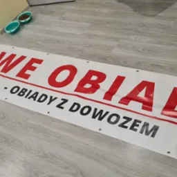Baner reklamowy z napisem 'Domowe Obiady - Codziennie od 13:00 - Obiady z dowozem od 20 zł' leżący na podłodze, obok miski dla zwierząt.