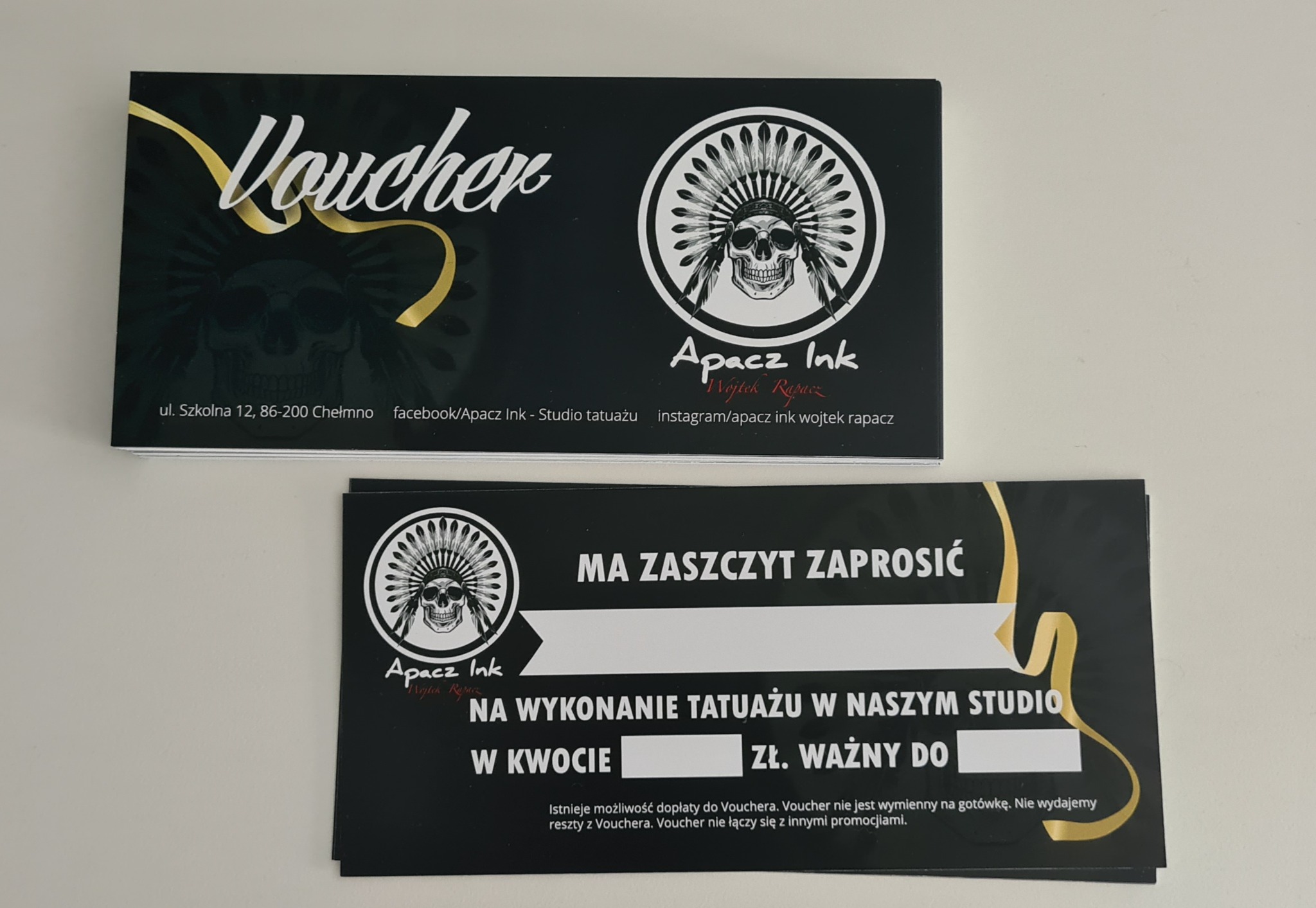 Dwa czarne vouchery z logo studia tatuażu Apacz Ink, z motywem czaszki w pióropuszu, zapraszające na wykonanie tatuażu.