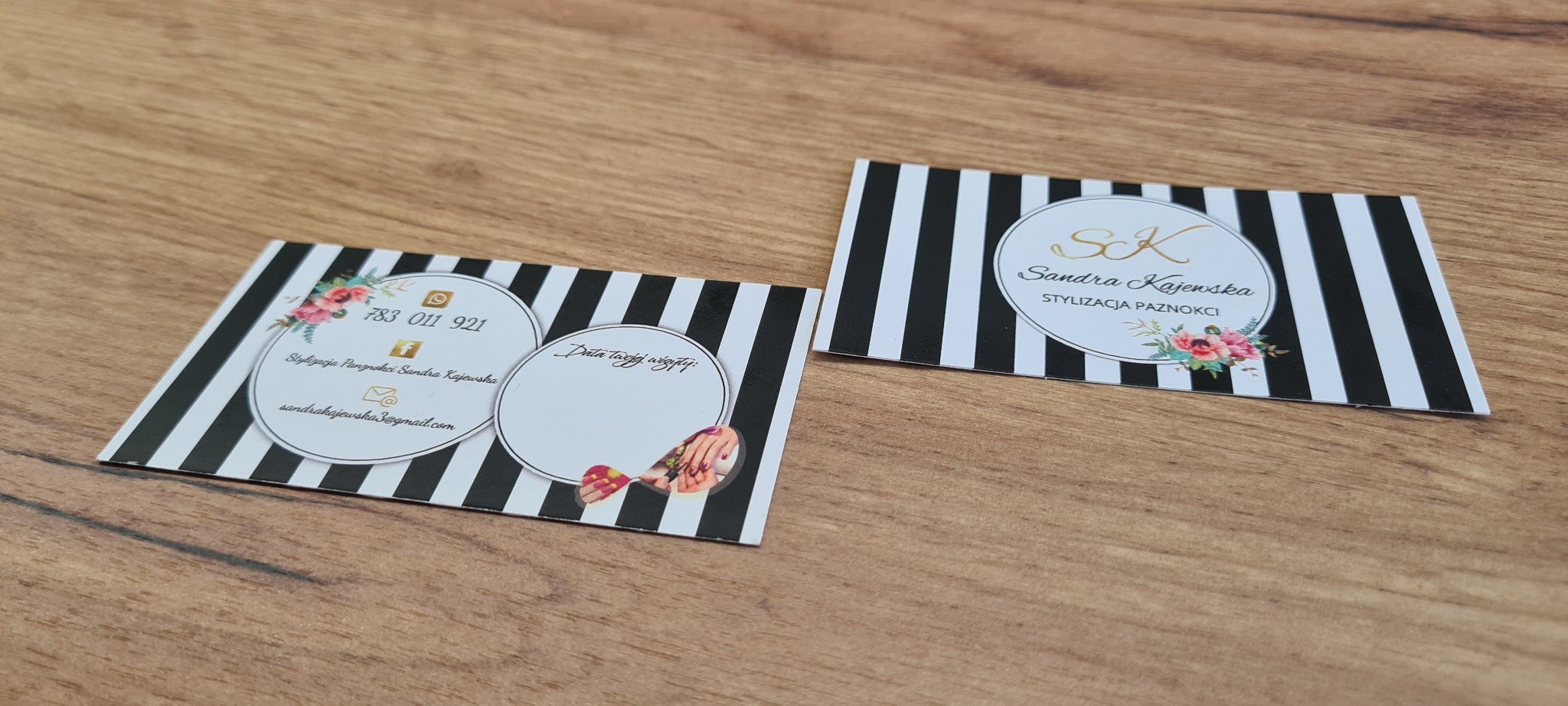 Dwie wizytówki z czarno-białymi paskami, kwiatowymi motywami i danymi kontaktowymi stylistki paznokci, leżące na drewnianym stole.
