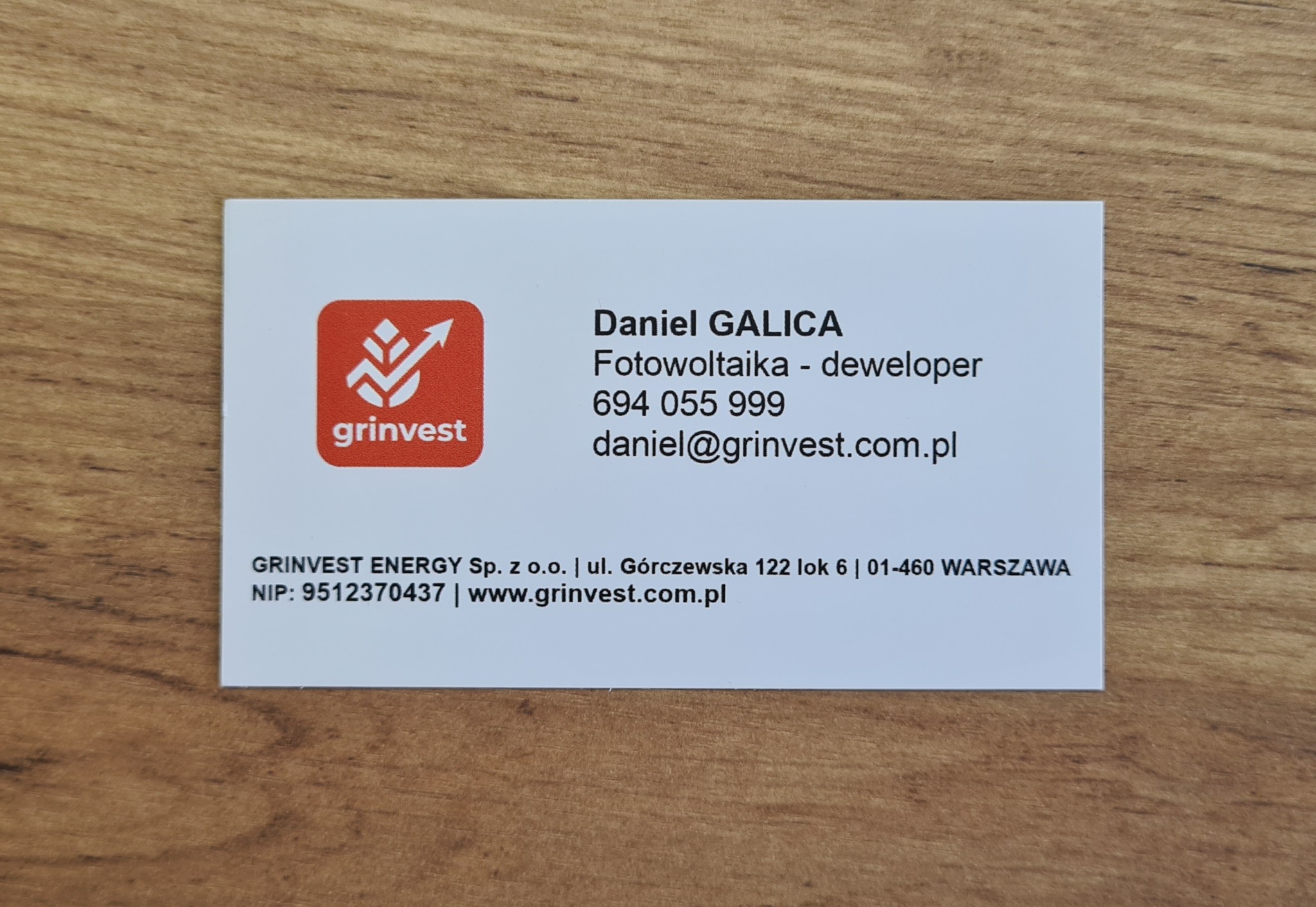 Wizytówka firmy Grinvest Energy z danymi kontaktowymi Daniela Galicy, specjalisty od fotowoltaiki, na tle drewnianego blatu.