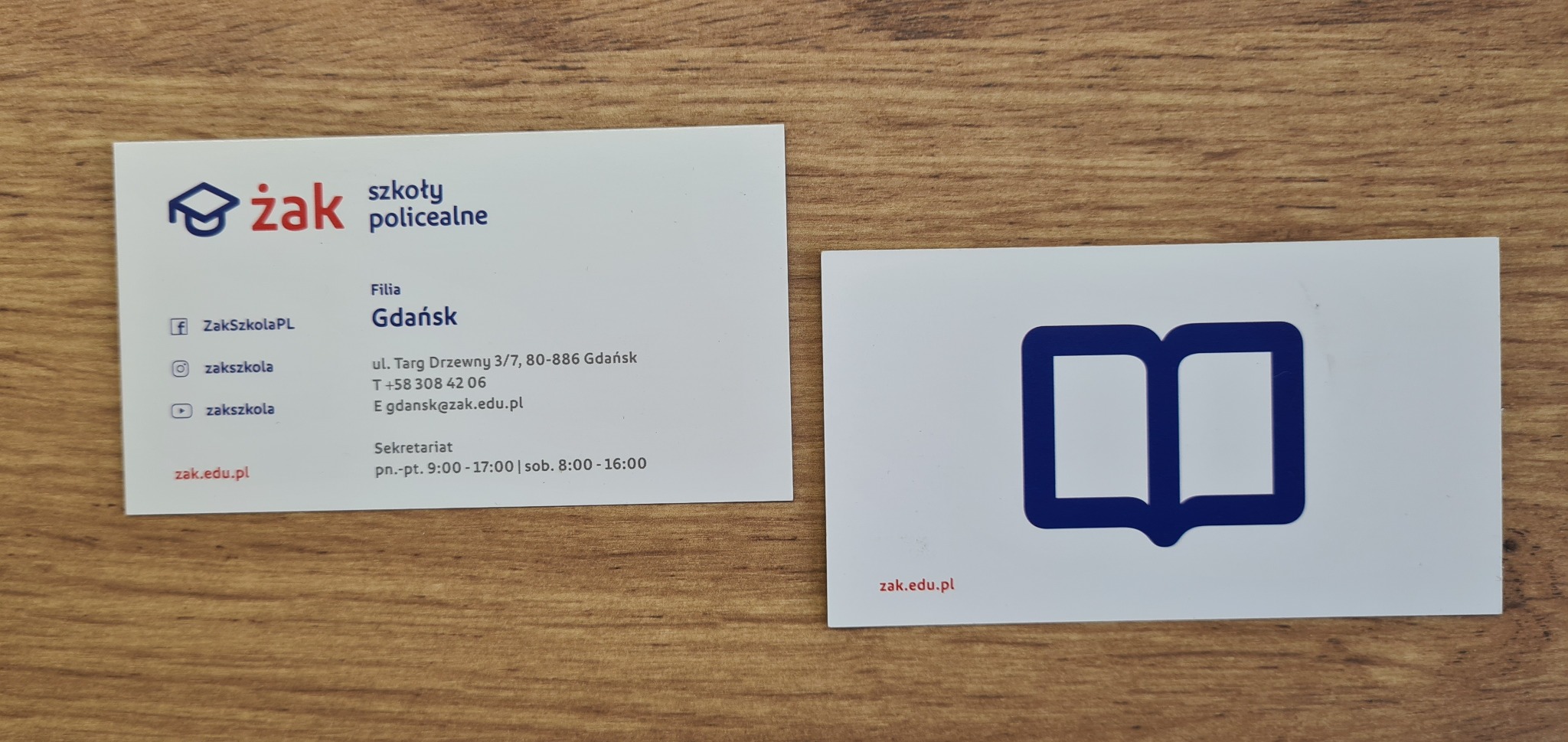 Dwie wizytówki firmy Żak Szkoły Policealne Gdańsk na drewnianym tle. Jedna wizytówka z logo i danymi kontaktowymi, druga z uproszczonym logo w postaci otwartej książki.