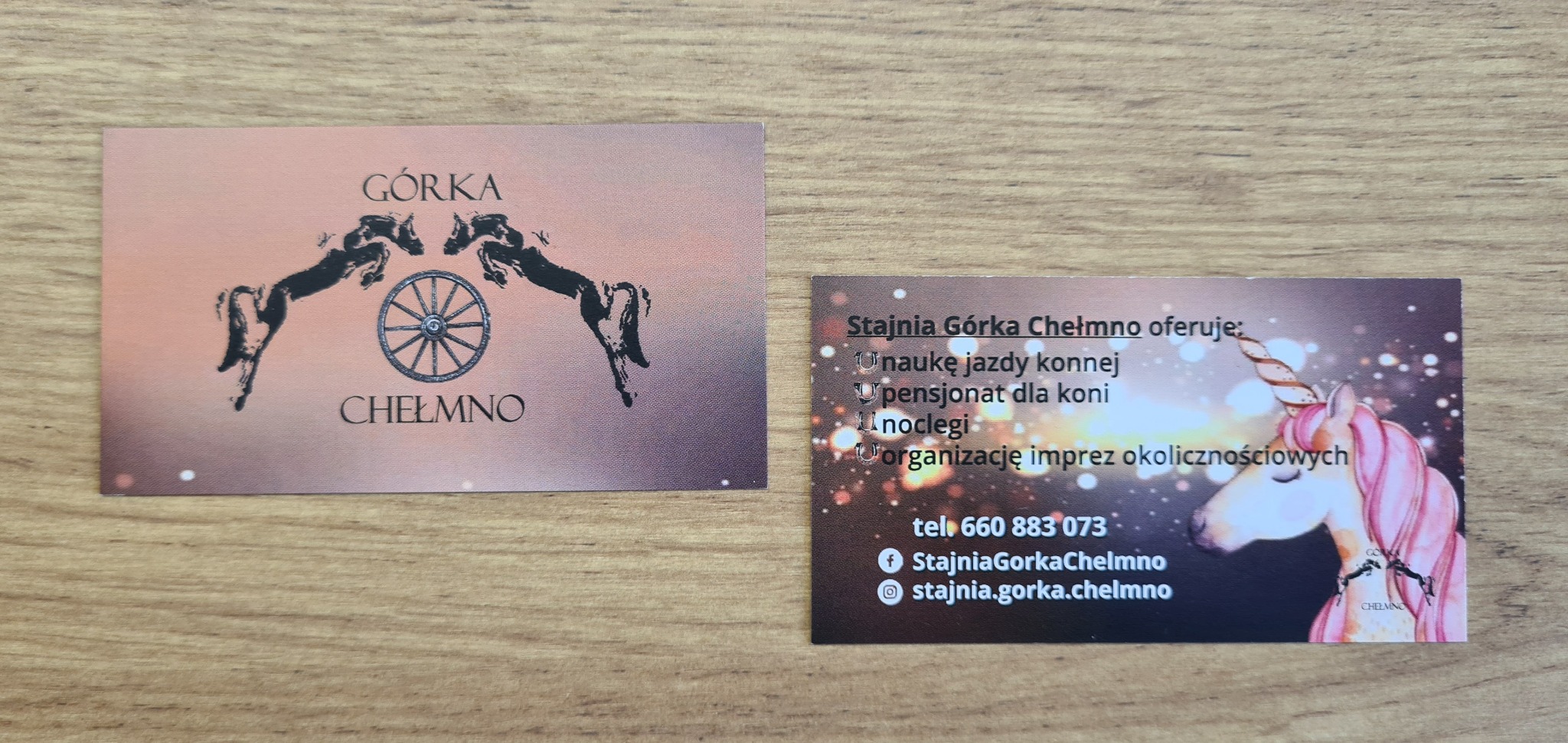 Dwie wizytówki na drewnianym tle. Jedna z grafiką koni i wozem, druga z jednorożcem, oferujące naukę jazdy konnej, pensjonat, noclegi i organizację imprez.