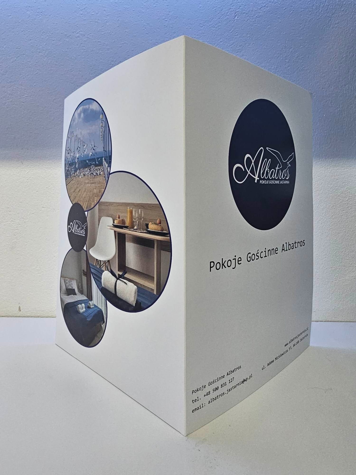 Trójwymiarowa, biała kostka z nadrukowanym logo i informacjami kontaktowymi Pokoi Gościnnych Albatros, zawierająca zdjęcia pokoi i otoczenia w okrągłych ramkach.