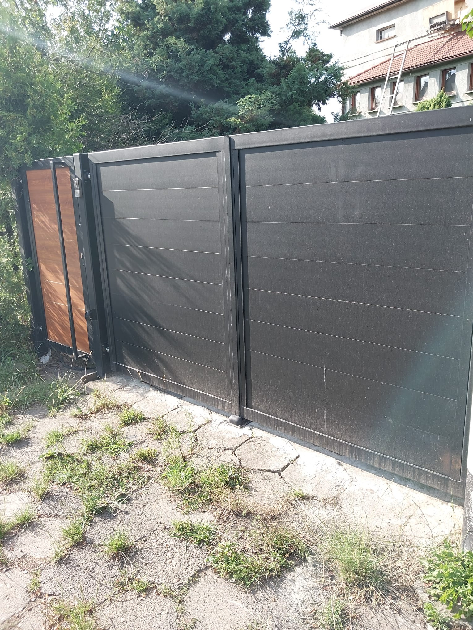 Nowoczesna brama wjazdowa z ciemnoszarymi, poziomymi panelami i elementami drewnopodobnymi, widoczna z bliska na tle zieleni i budynku.