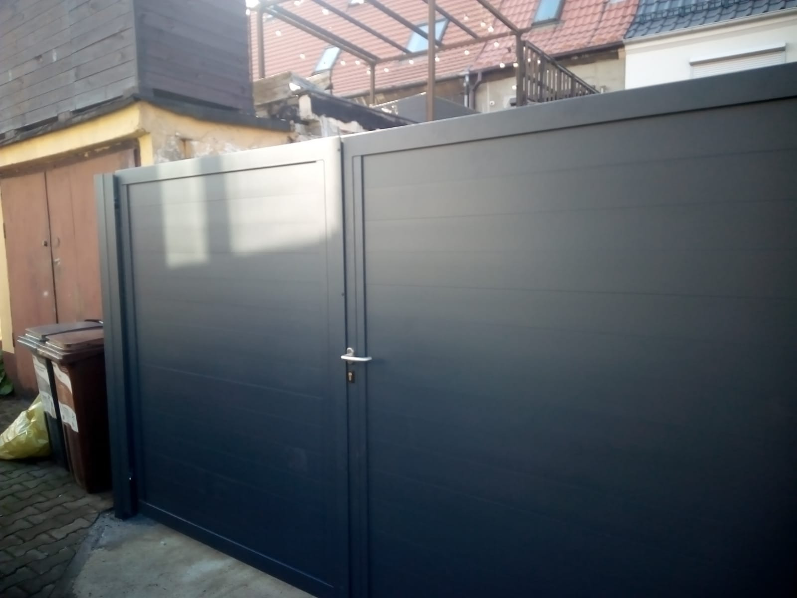 Nowoczesna, antracytowa brama wjazdowa z poziomymi panelami, widoczna z zewnątrz posesji, z brązowym garażem i koszami na śmieci w tle.