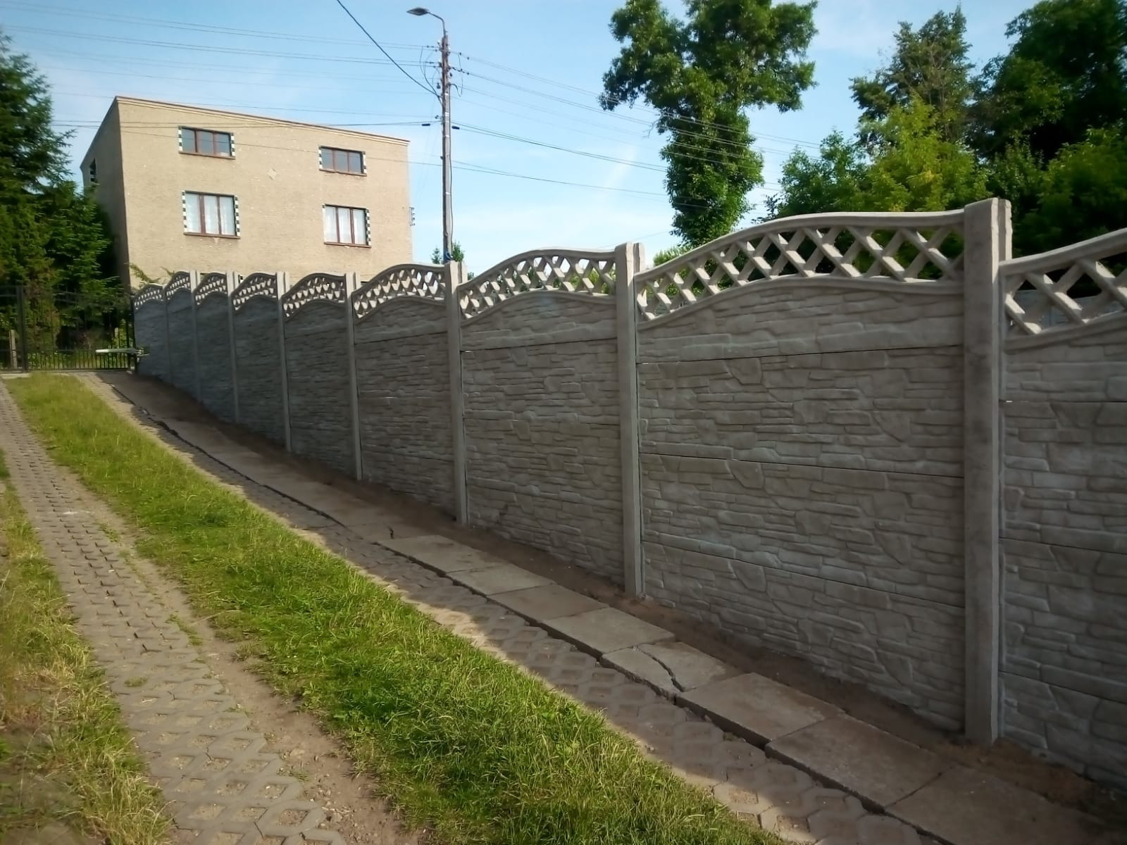 Długi, szary, betonowy płot z dekoracyjnym, ażurowym wykończeniem górnej części, oddzielający posesję od drogi z betonowej kostki, w tle budynek mieszkalny i drzewa.