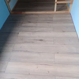 Ułożone panele podłogowe w jasnym odcieniu drewna, widoczne fragmenty niebieskiej ściany i drewnianej konstrukcji pod sufitem.