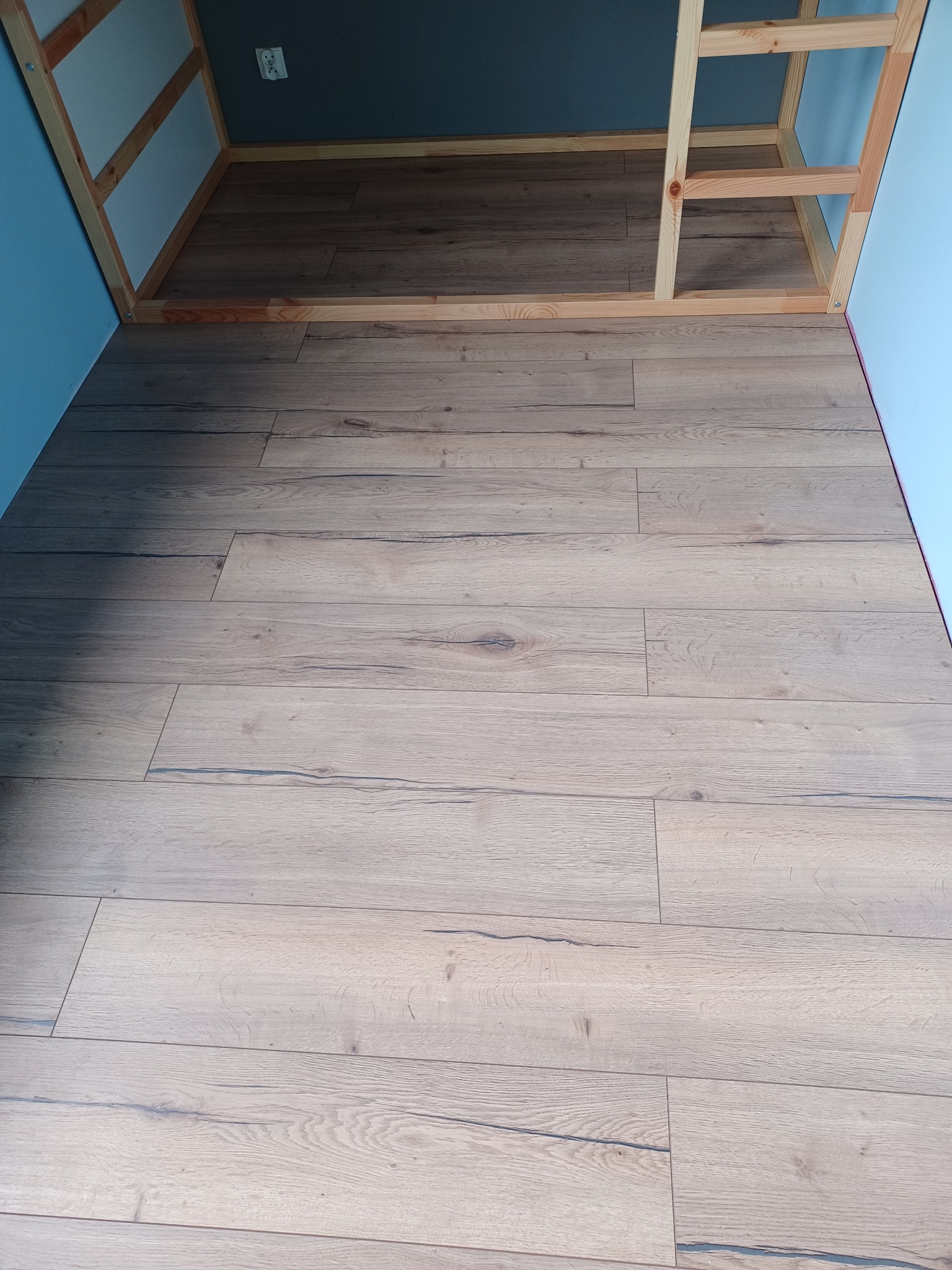 Ułożone panele podłogowe w jasnym odcieniu drewna, widoczne fragmenty niebieskiej ściany i drewnianej konstrukcji pod sufitem.