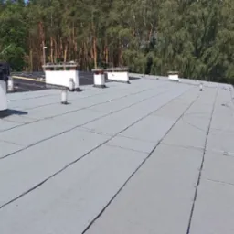 Płaski dach pokryty szarą papą termozgrzewalną z białymi kominami i elementami wentylacyjnymi, na czarnej papie leżą rolki papy i narzędzia dekarskie, w tle drzewa i budynki gospodarcze.