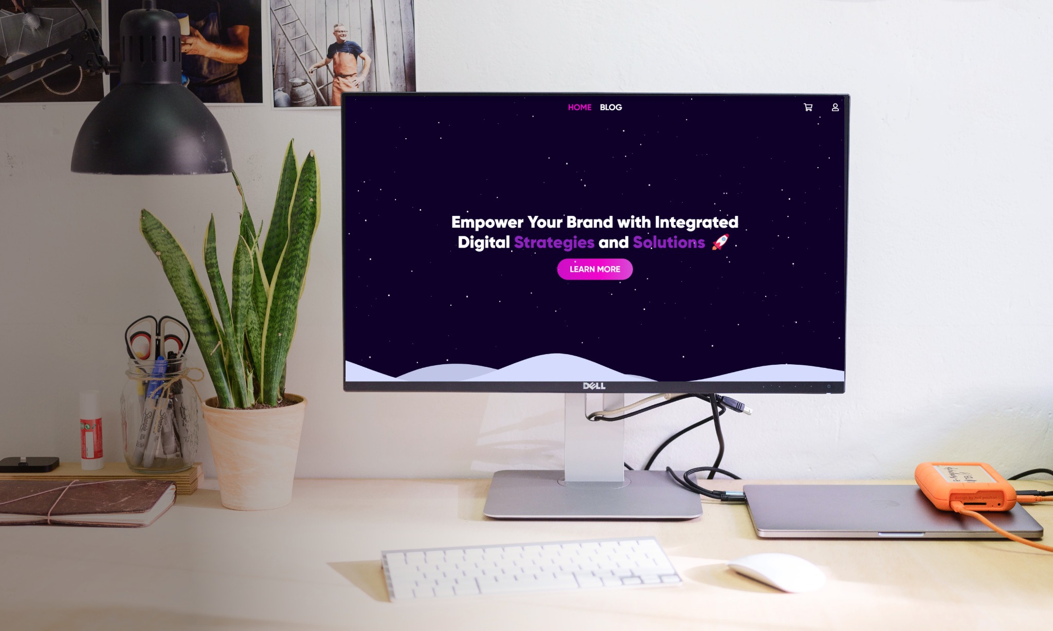 Monitor Dell wyświetlający stronę internetową z motywem kosmicznym i hasłem 'Empower Your Brand with Integrated Digital Strategies and Solutions' na biurku z klawiaturą, myszką, laptopem i rośliną...