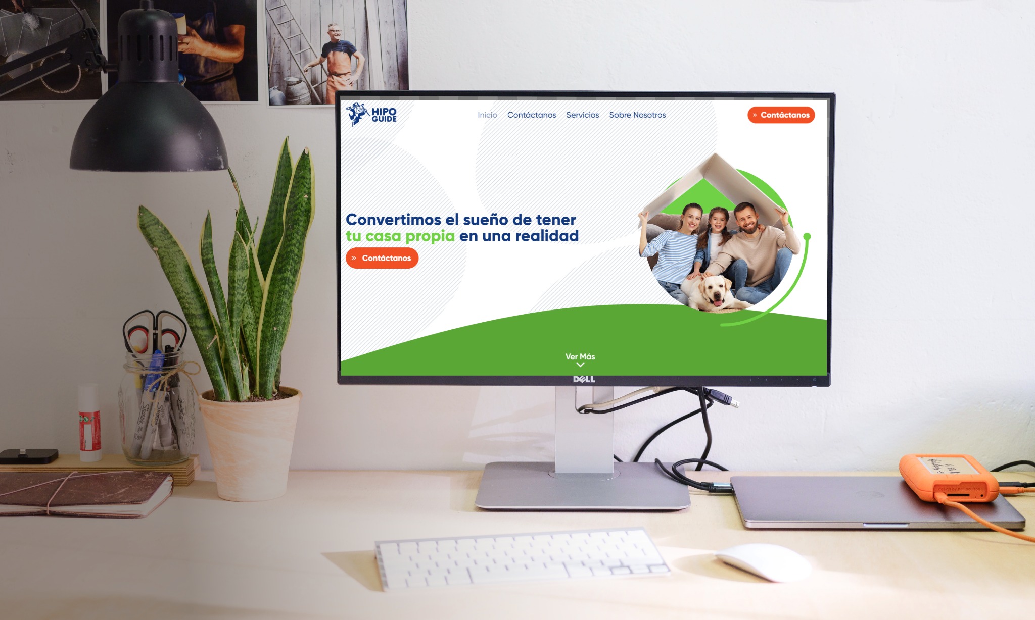 Monitor Dell wyświetlający stronę internetową z hasłem 'Convertimos el sueño de tener tu casa propia en una realidad', biurko z lampką, rośliną w doniczce i akcesoriami biurowymi w tle.