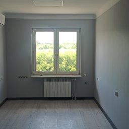 Łukasz Stępień - Pomalowane na szaro wnętrze pokoju z panelami podłogowymi, grzejnikiem pod oknem i klimatyzacją na ścianie.